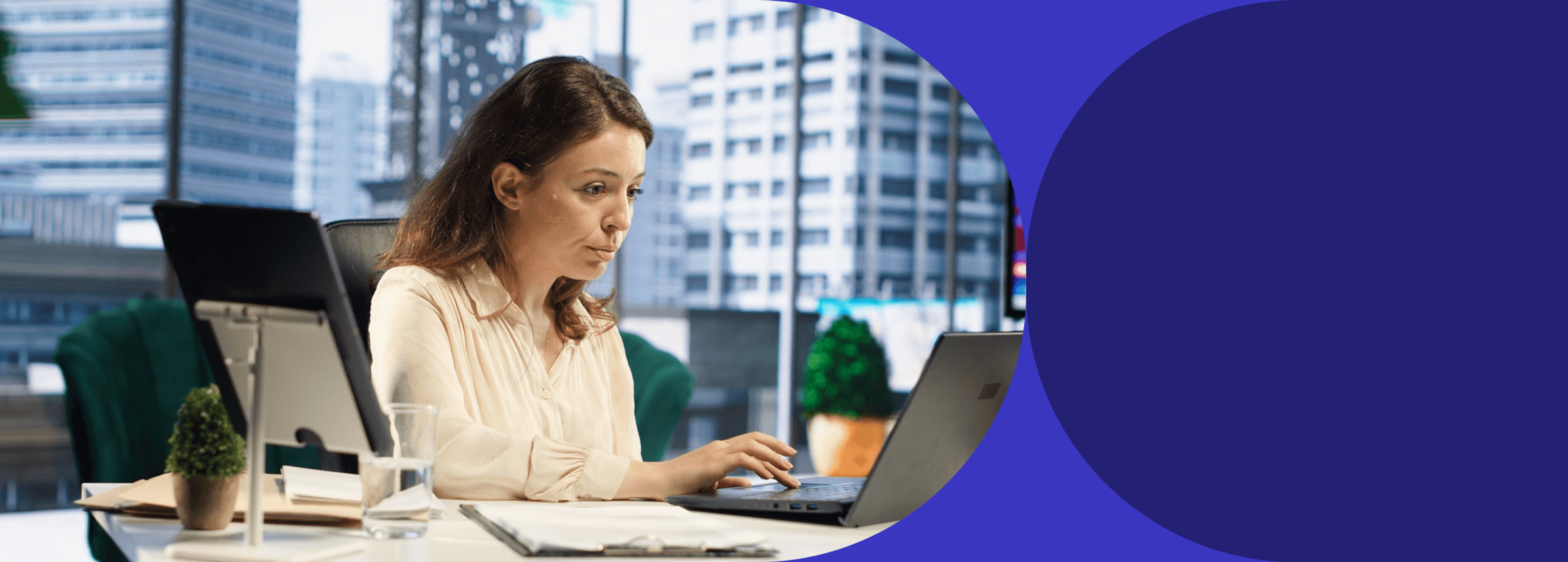 Woman typing on a laptop at an office desk, city skyline in background.