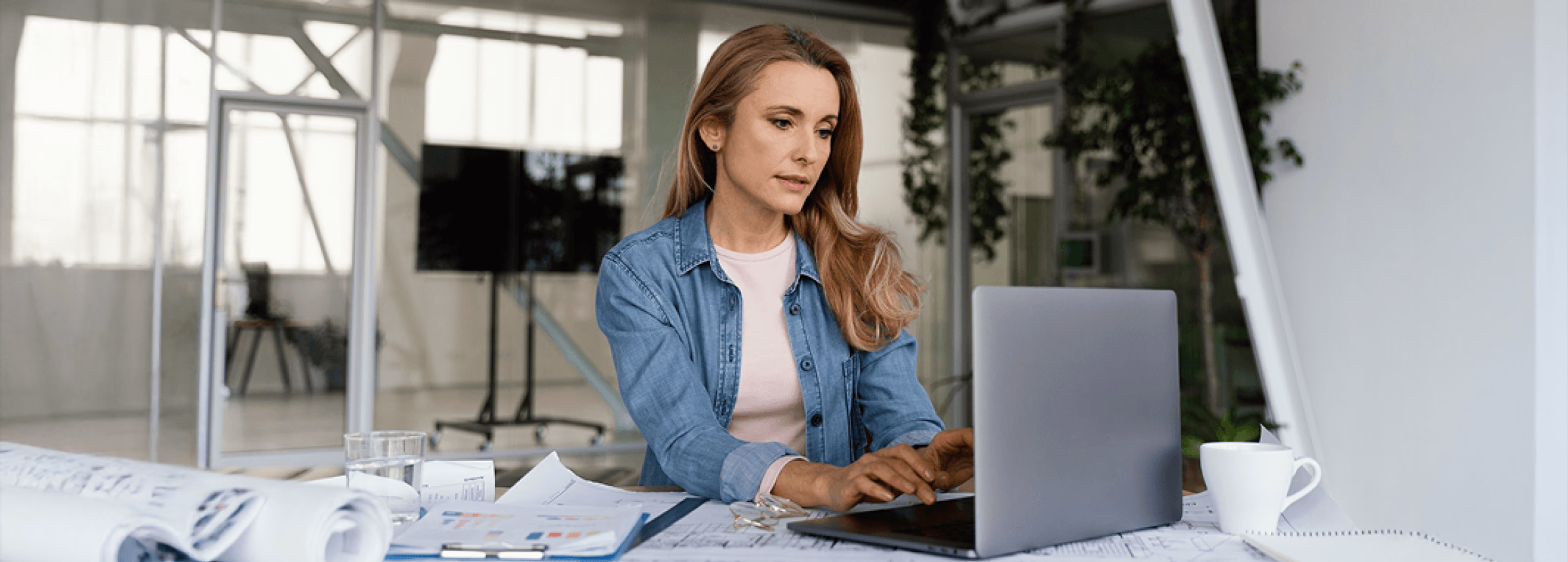 A woman working on a laptop at a modern office desk with blueprints, documents, and a coffee mug in front of her.