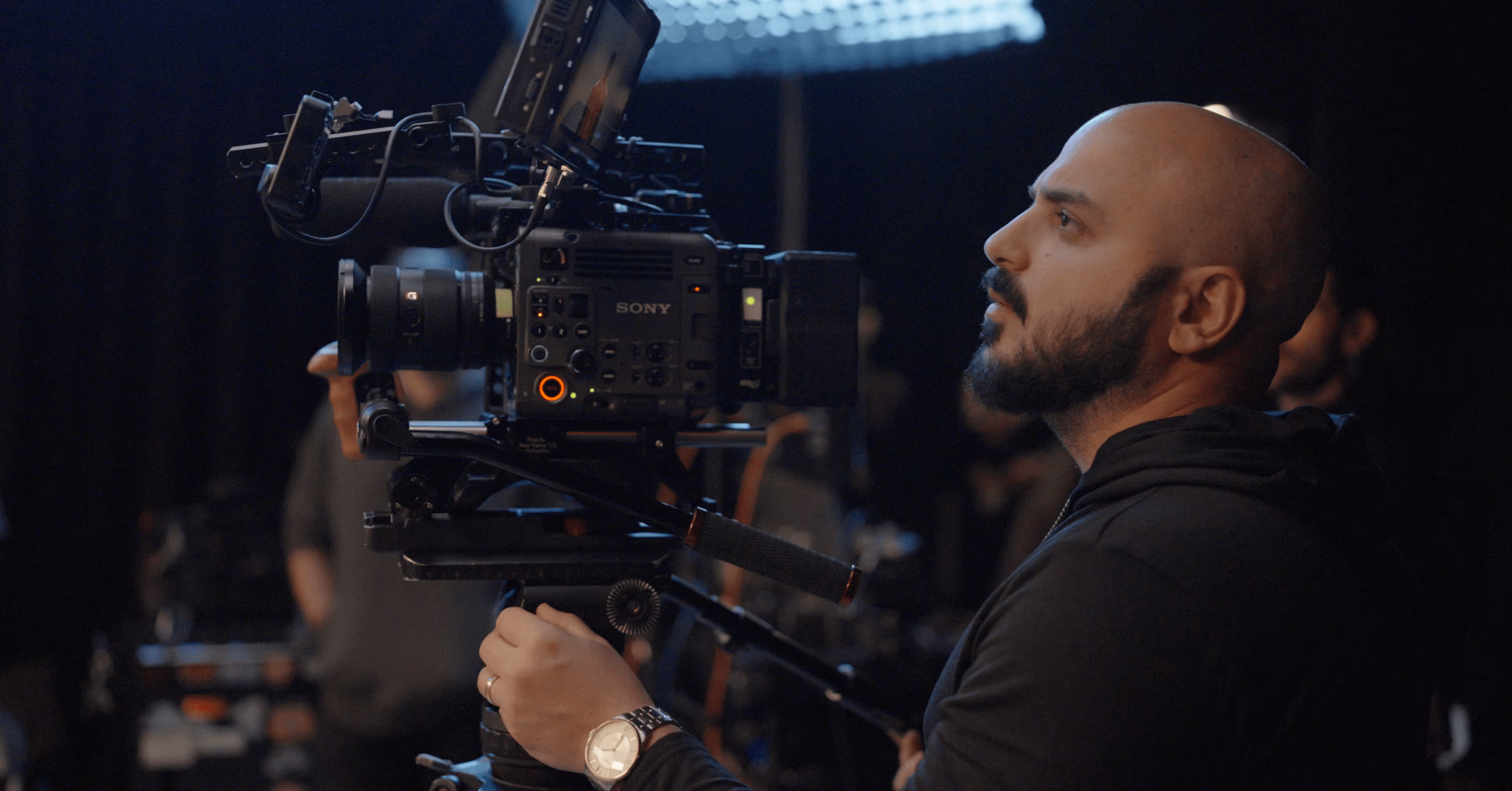 A close-up profile shot of a cinematographer operating a professional Sony cinema camera on a dimly lit production set.