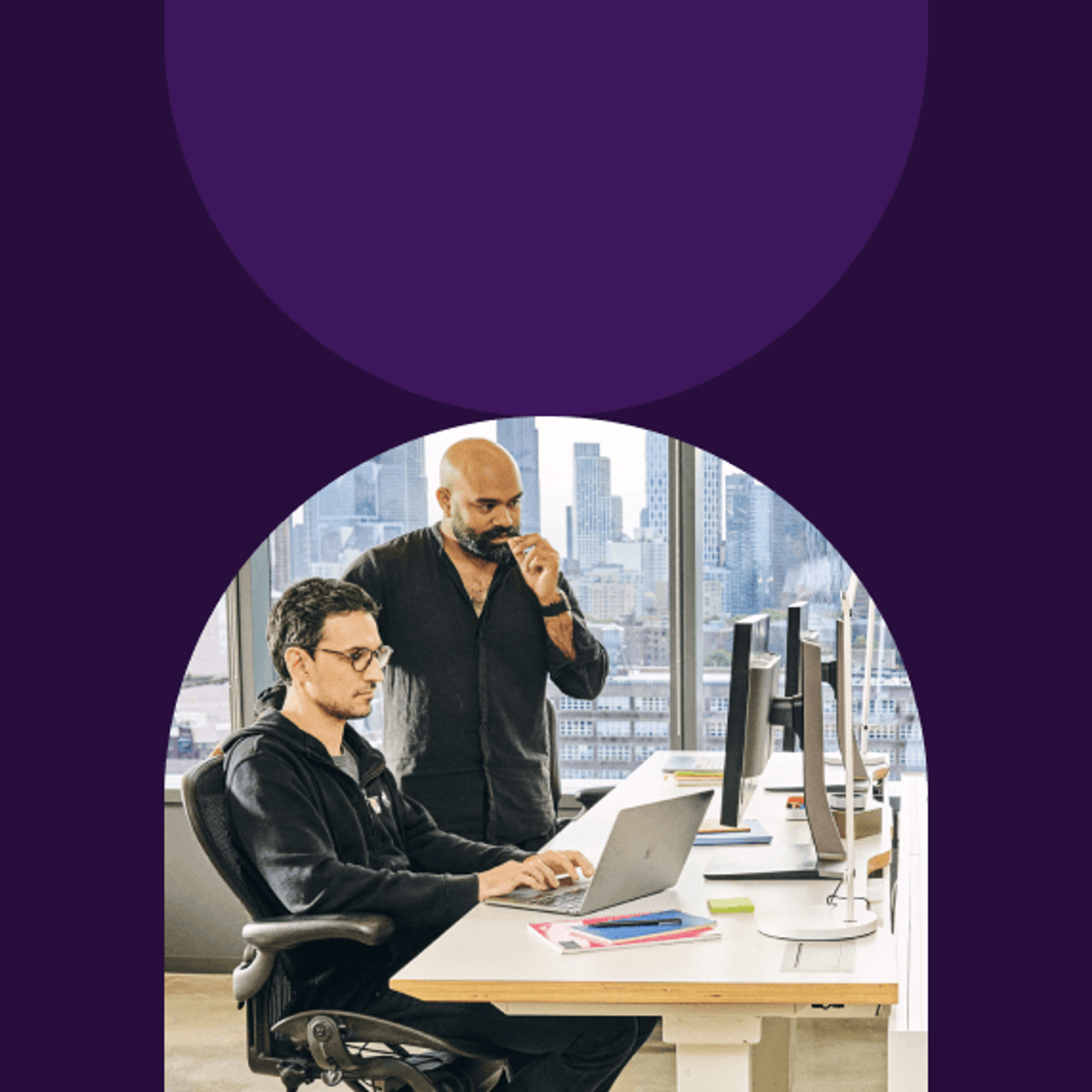 Two creative professionals collaborating at a modern office desk with laptops and monitors.